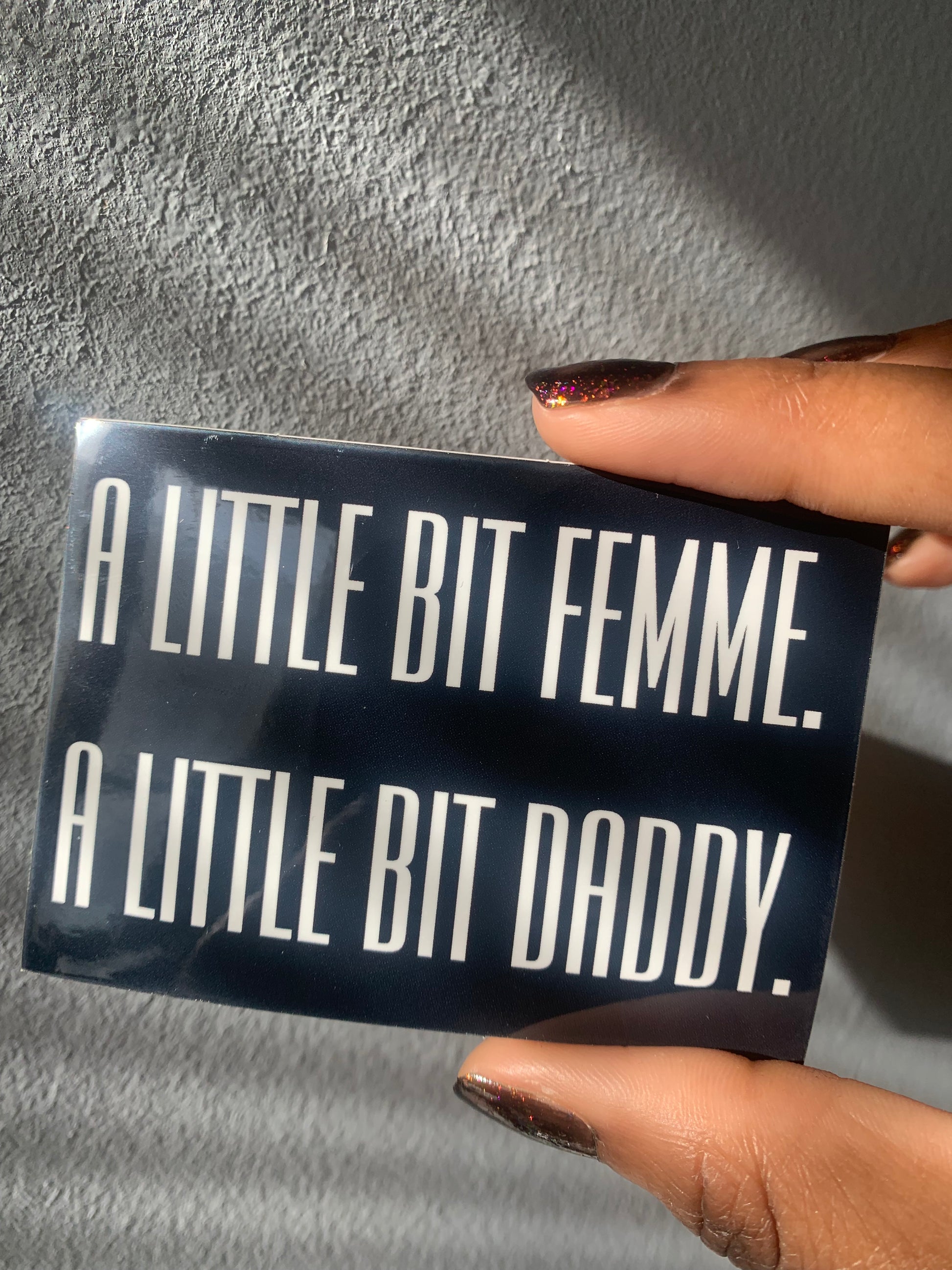 A rectangular black and white glossy vinyl sticker held between two fingers that reads A LITTLE BIT FEMME. A LITTLE BIT DADDY. In bold white text over a black background.