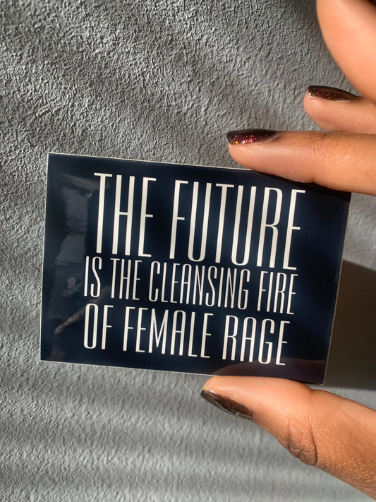 A rectangular black and white glossy vinyl sticker held between two fingers that reads THE FUTURE IS THE CLEANSING FIRE OF FEMALE RAGE in bold white text over a black background.