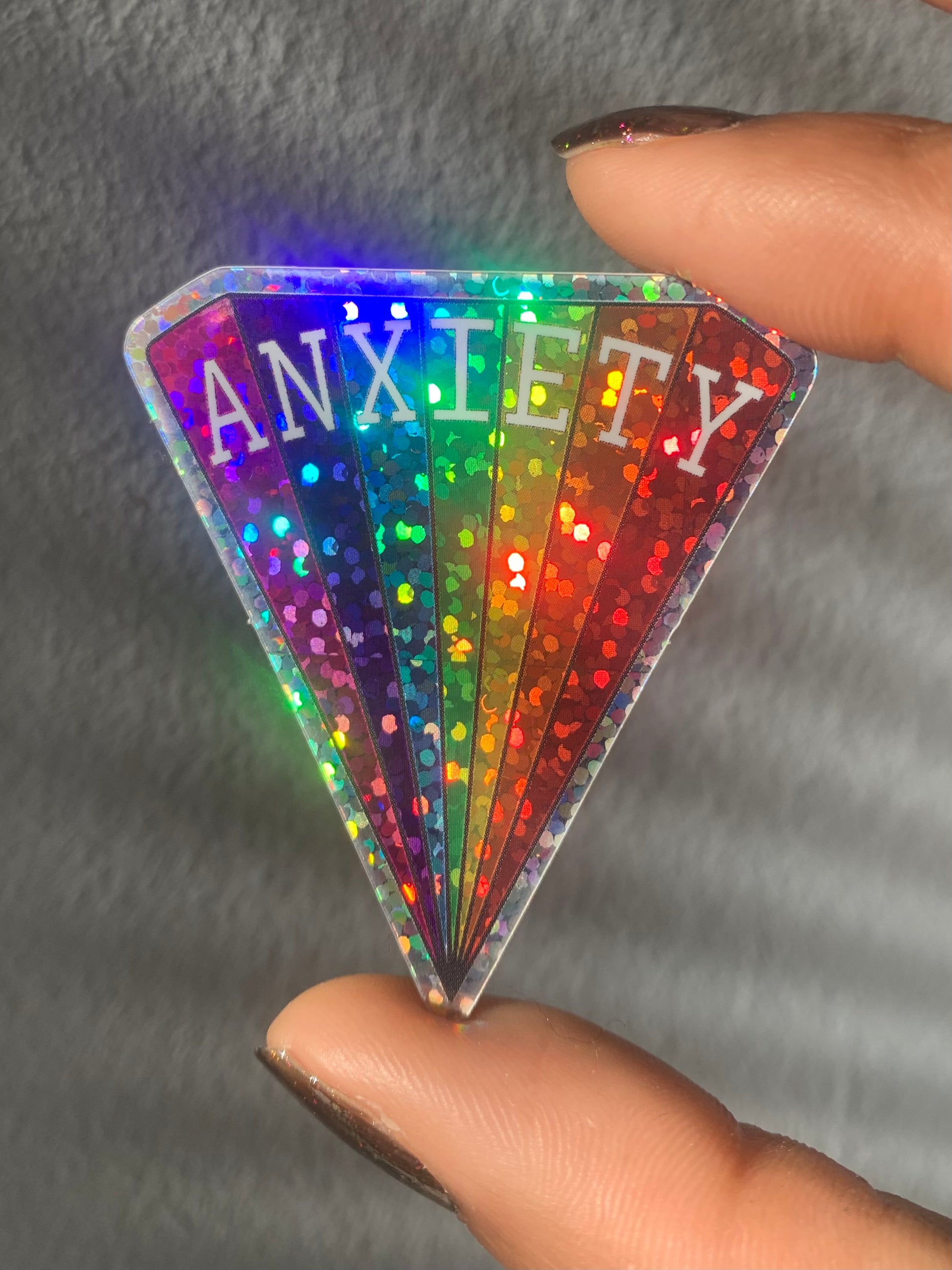 Triangular vinyl sticker with a rainbow design in glitter that reads ANXIETY across the top, measuring 2 x 1.8 inches being held between two fingers.