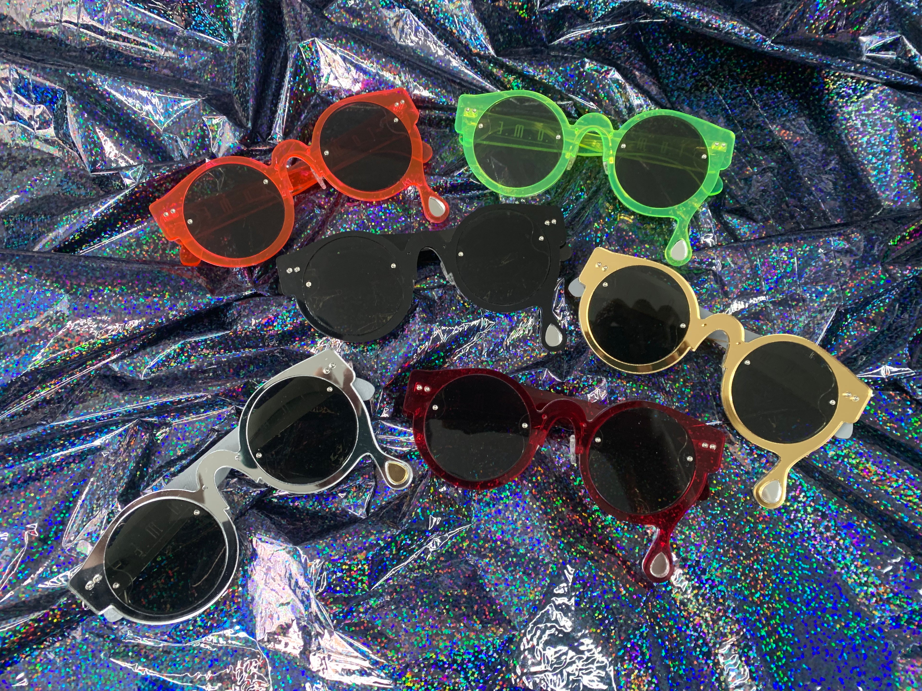A assortment of acrylic laser cut sunglasses with a teardrop accent dripping down the right side of the glasses. Different colors shown are neon pink, neon green, black, transparent red, silver mirror, and gold mirror.