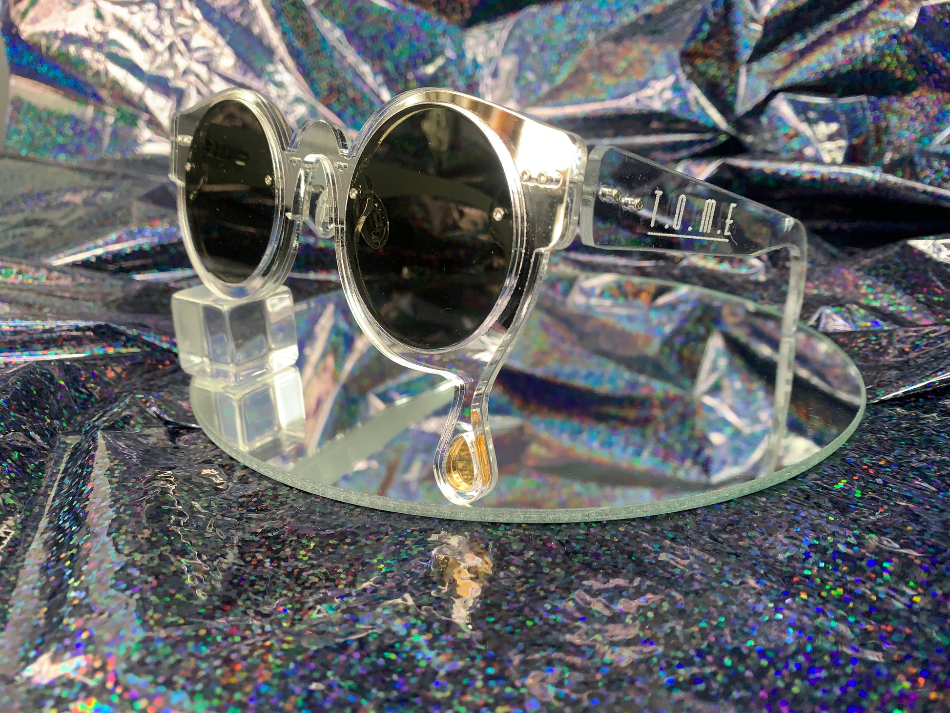 A side view of acrylic laser cut sunglasses with a teardrop accent dripping down the right side of the glasses with the brand logo T.O.M.E. etched on the temple. The color of the sunglasses shown are silver mirror.