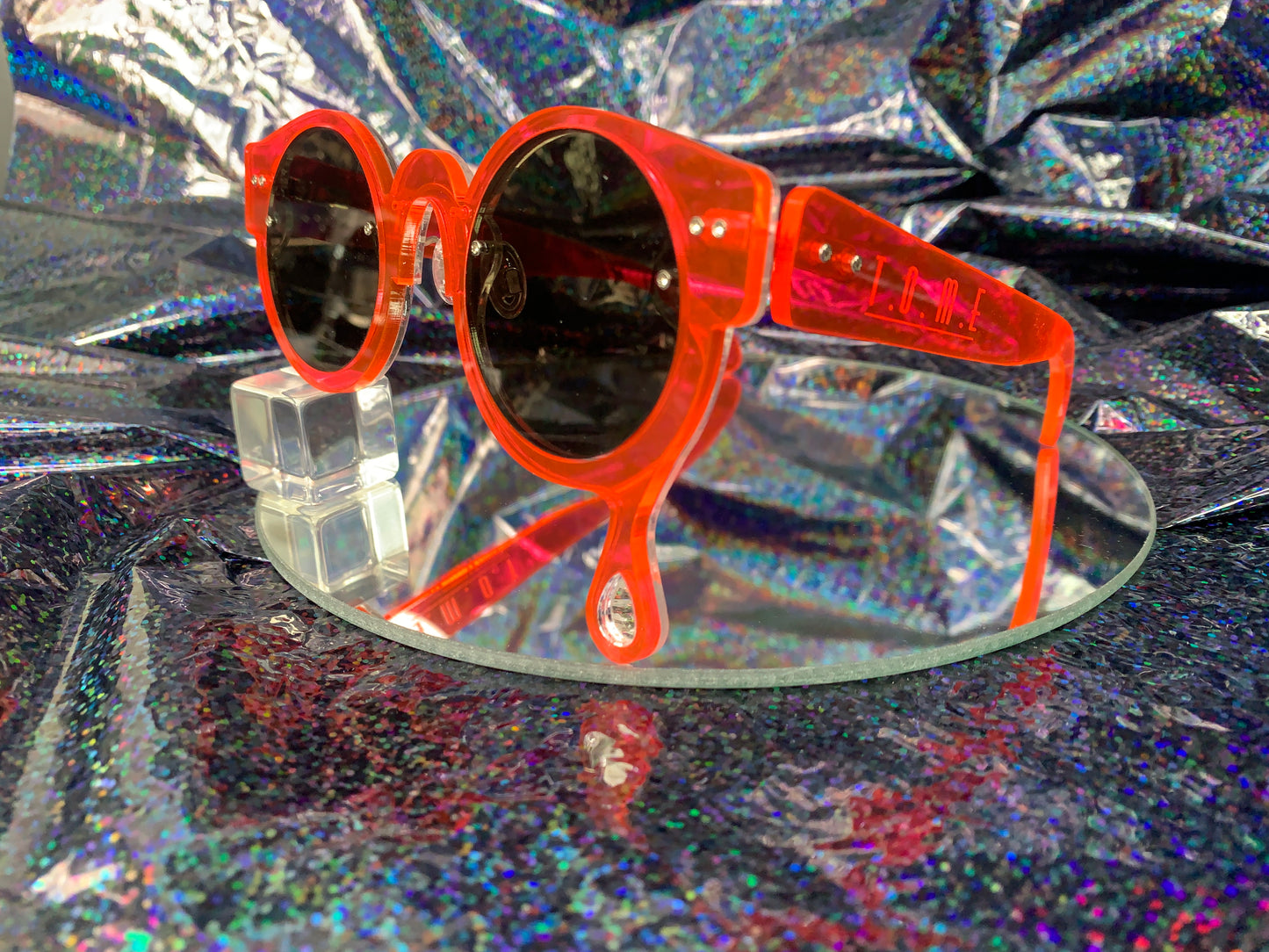 A side view of acrylic laser cut sunglasses with a teardrop accent dripping down the right side of the glasses with the brand logo T.O.M.E. etched on the temple. The color of the sunglasses shown are neon pink.