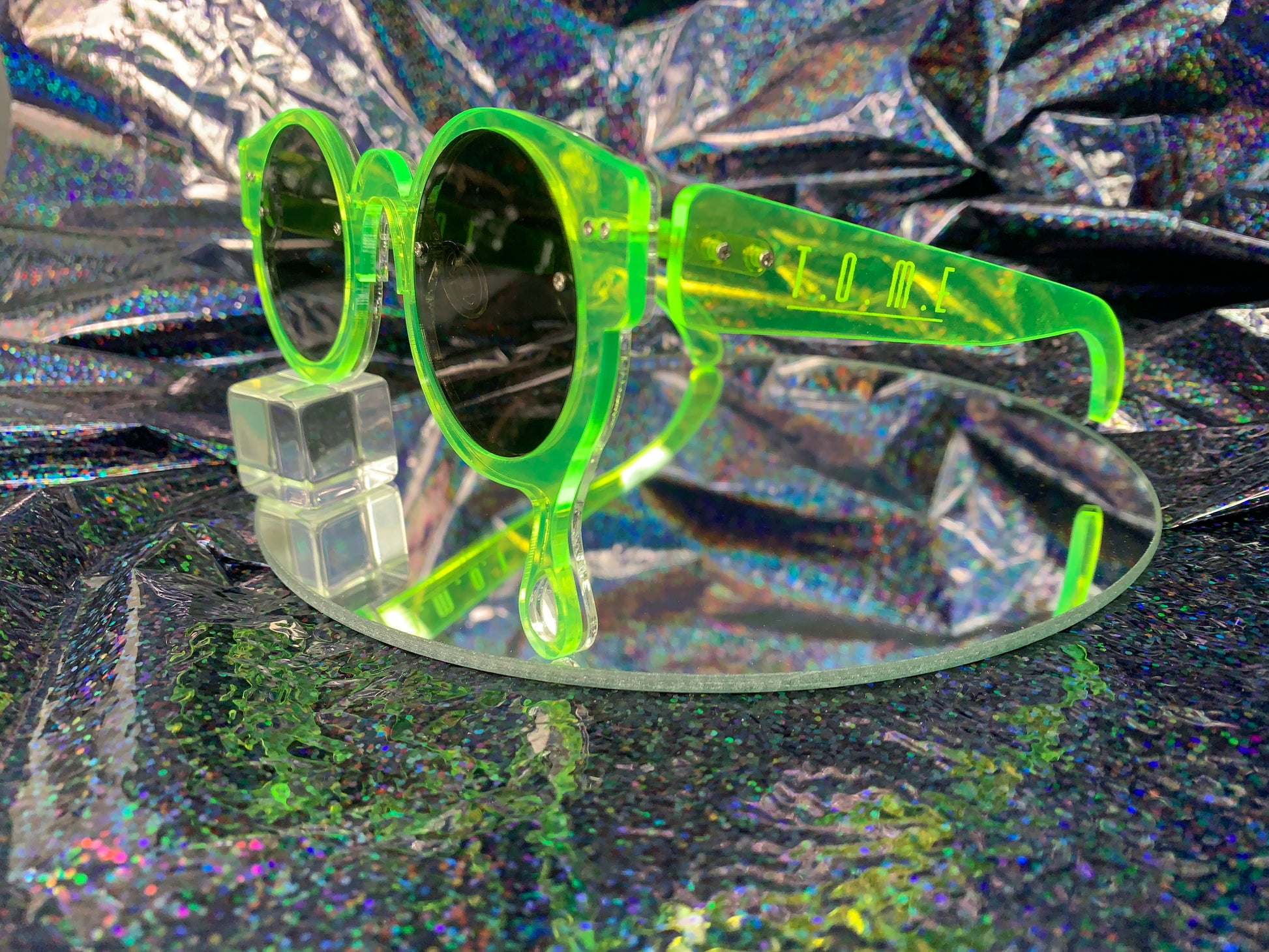 A side view of acrylic laser cut sunglasses with a teardrop accent dripping down the right side of the glasses with the brand logo T.O.M.E. etched on the temple. The color of the sunglasses shown are neon green.