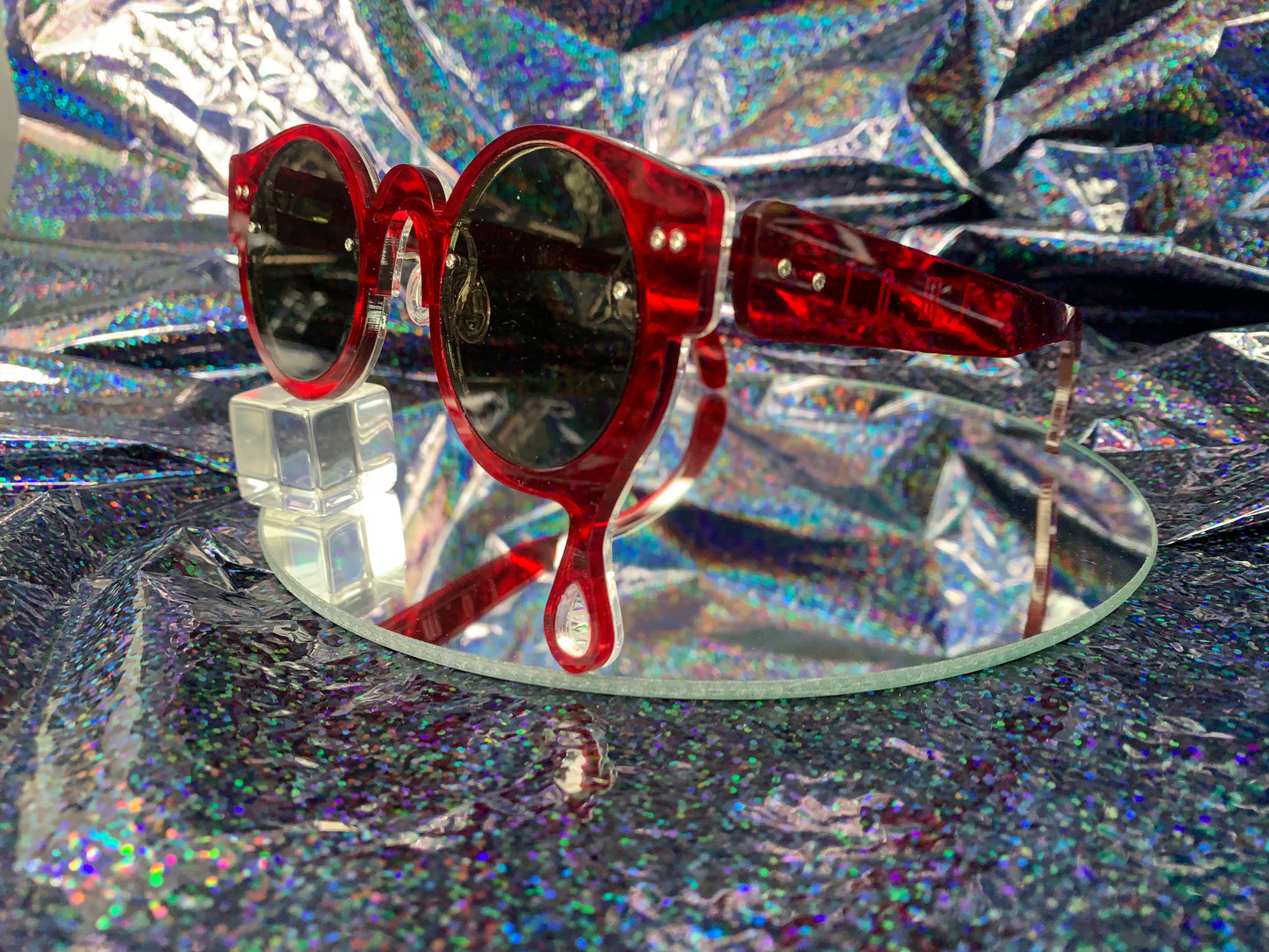 A side view of acrylic laser cut sunglasses with a teardrop accent dripping down the right side of the glasses with the brand logo T.O.M.E. etched on the temple. The color of the sunglasses shown are transparent red.