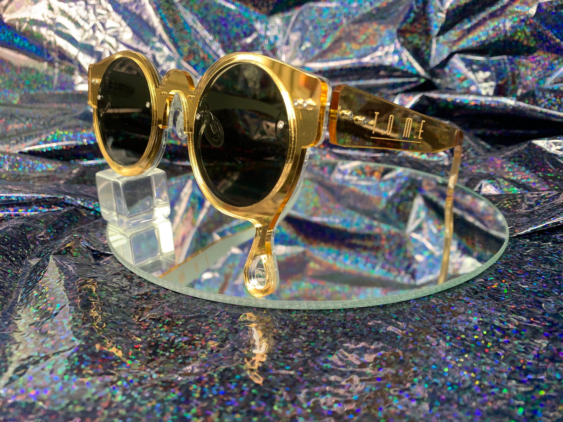 A side view of acrylic laser cut sunglasses with a teardrop accent dripping down the right side of the glasses with the brand logo T.O.M.E. etched on the temple. The color of the sunglasses shown are gold mirror.
