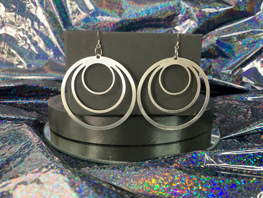 A pair of round stainless steel earrings measuring 2.5" hanging in front of a rainbow glitter background.