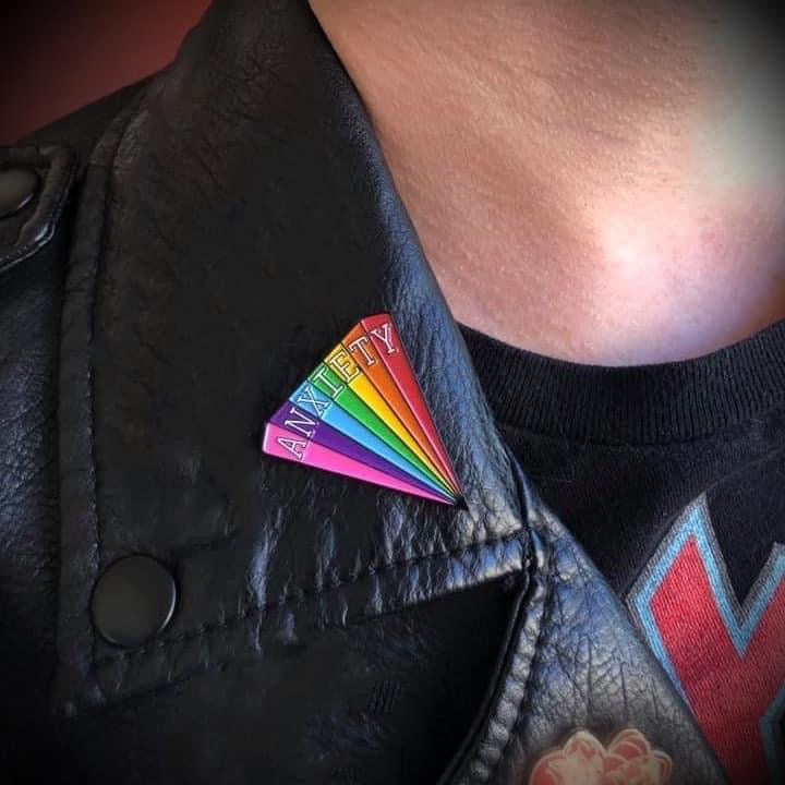 Close up on the collar of a black leather jacket with a triangular enamel pin pinned to it that reads ANXIETY in white text across it..