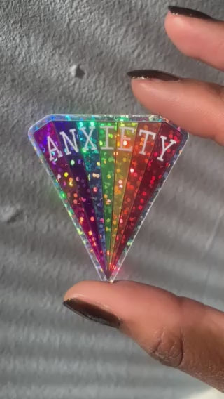 4 second movie of a triangular vinyl sticker with a rainbow design in glitter that reads ANXIETY across the top, measuring 2 x 1.8 inches being held between two fingers showing the effect of the glitter in sunlight..