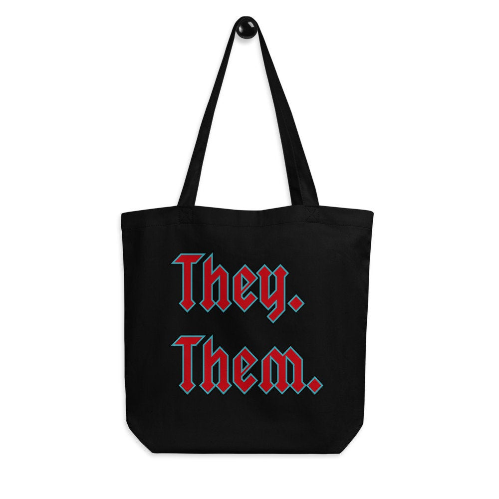 Black tote bag that reads THEY. THEM. in red and aqua blue text, on a white background 