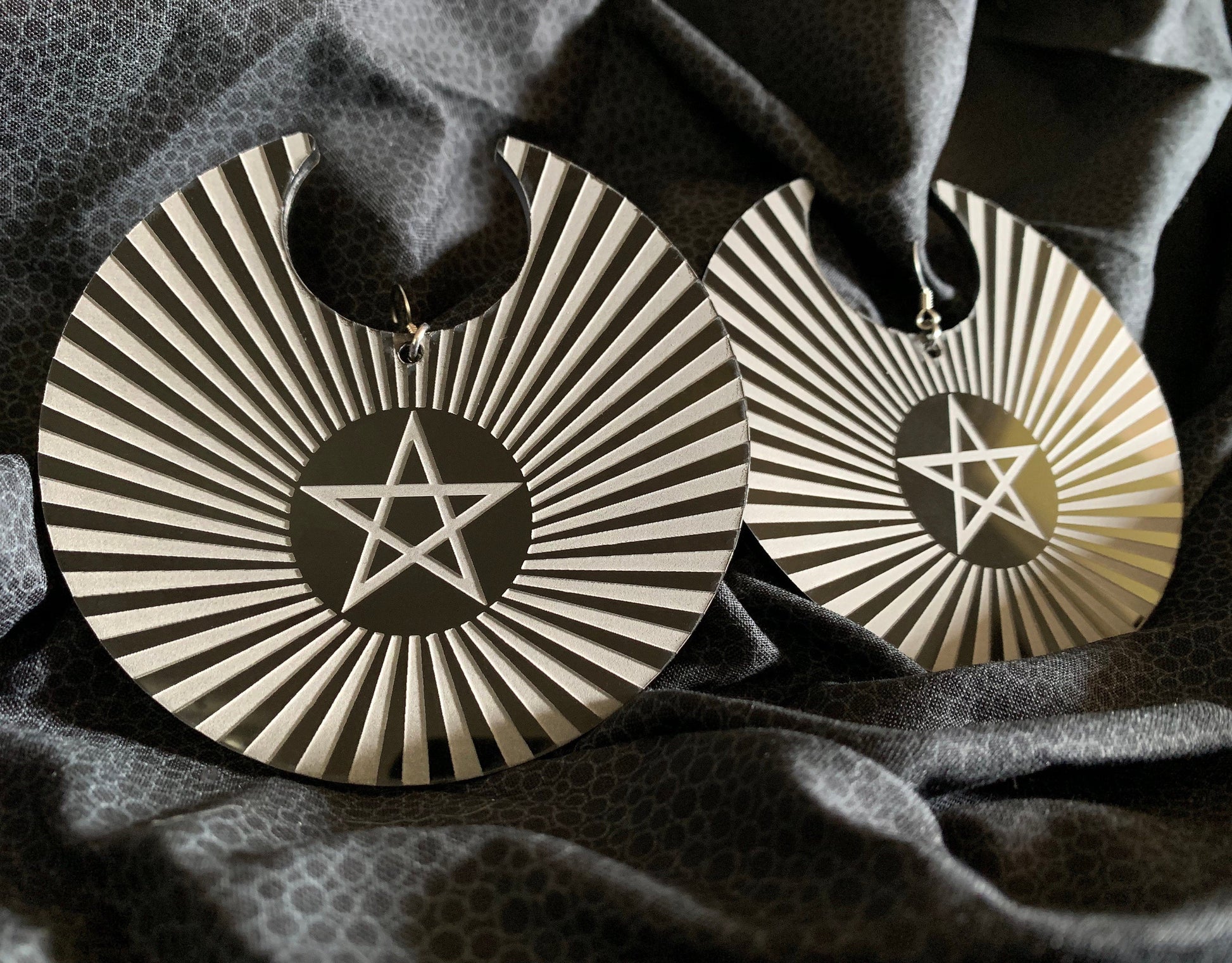 Round black and white acrylic earrings with a pentacle etched into the center surrounded by a sunbeam pattern filling the entirety of the earrings.